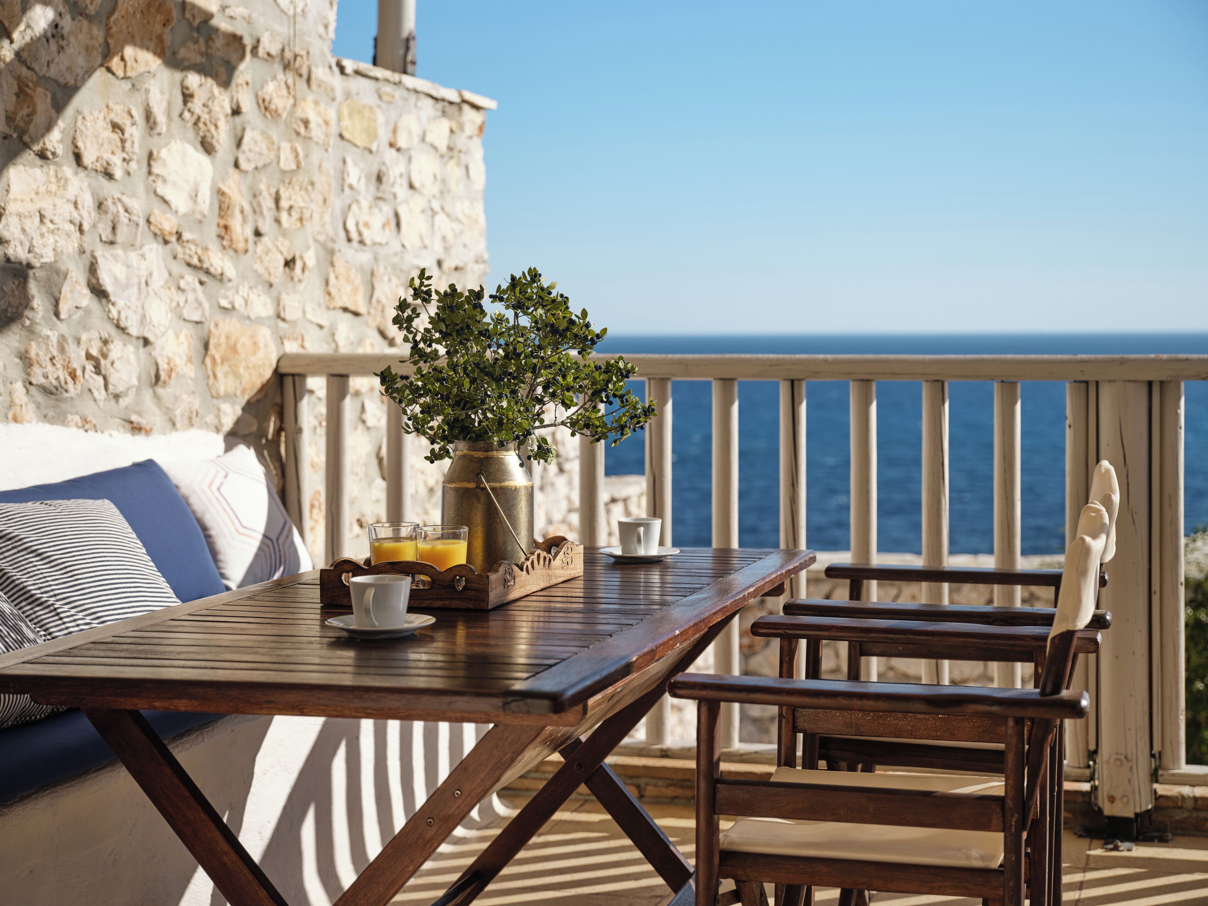 Ein Holztisch mit einer Vase voller Grünpflanzen, zwei Gläser Orangensaft und einer Tasse auf einem Balkon mit Blick auf das Meer.