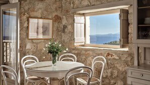 A stone-walled room with a round table and white chairs, a glass vase with flowers, and a view of the sea through a window.