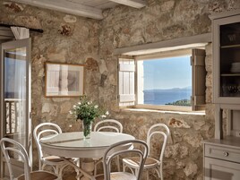 A stone-walled room with a round table and white chairs, a glass vase with flowers, and a view of the sea through a window.