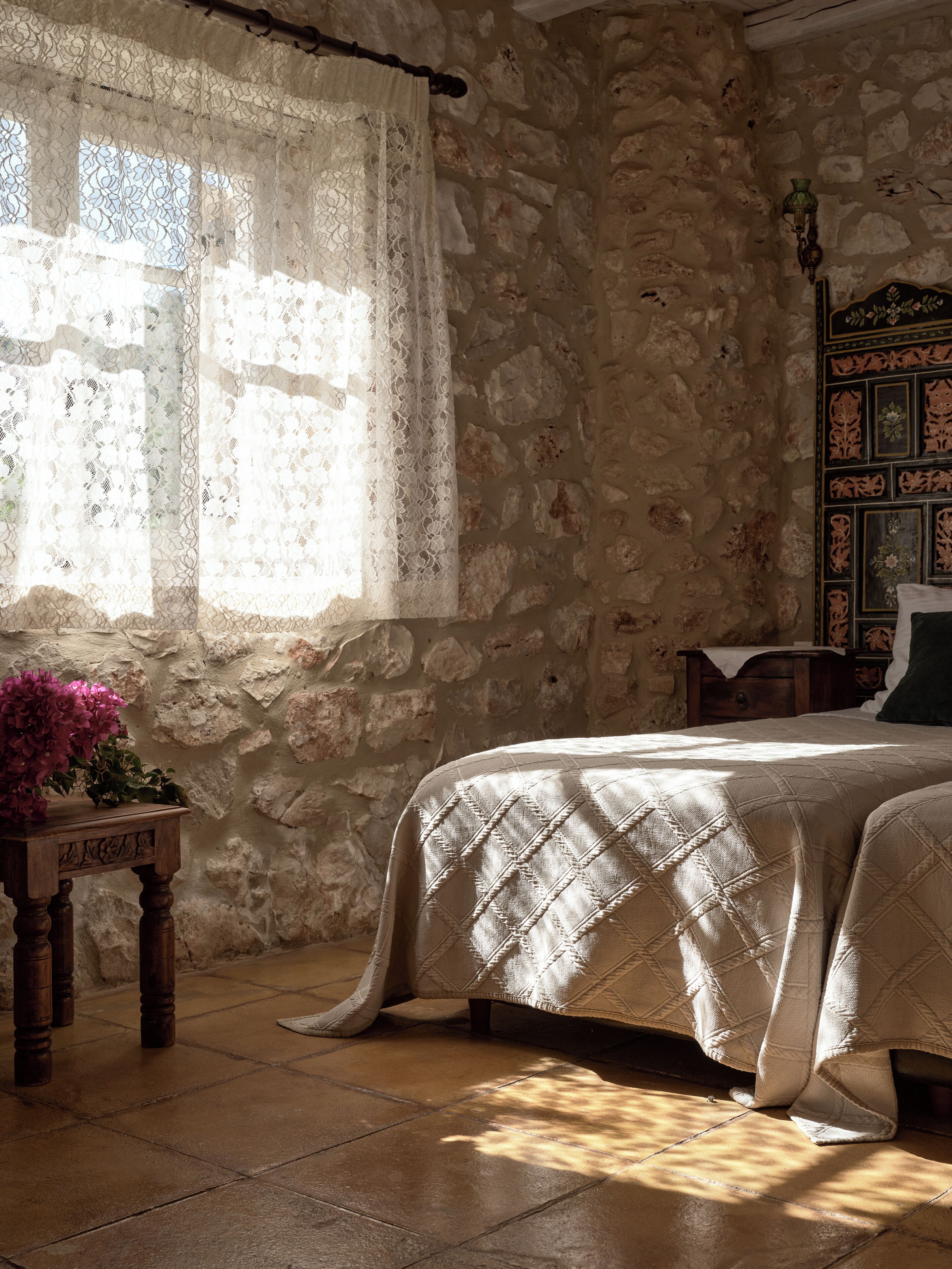 Un dormitorio con pared de piedra, una cama con colcha, una mesita de noche de madera con un florero y una ventana con cortinas de encaje.