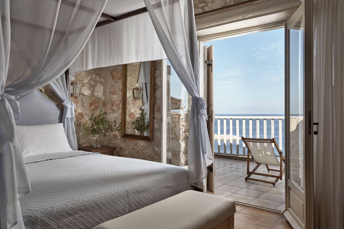 A bedroom with a large bed, a bench, and a balcony with a view of the sea.