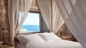 A bedroom with a bed, white curtains, a nightstand with a lamp, and a view of the sea through a window.