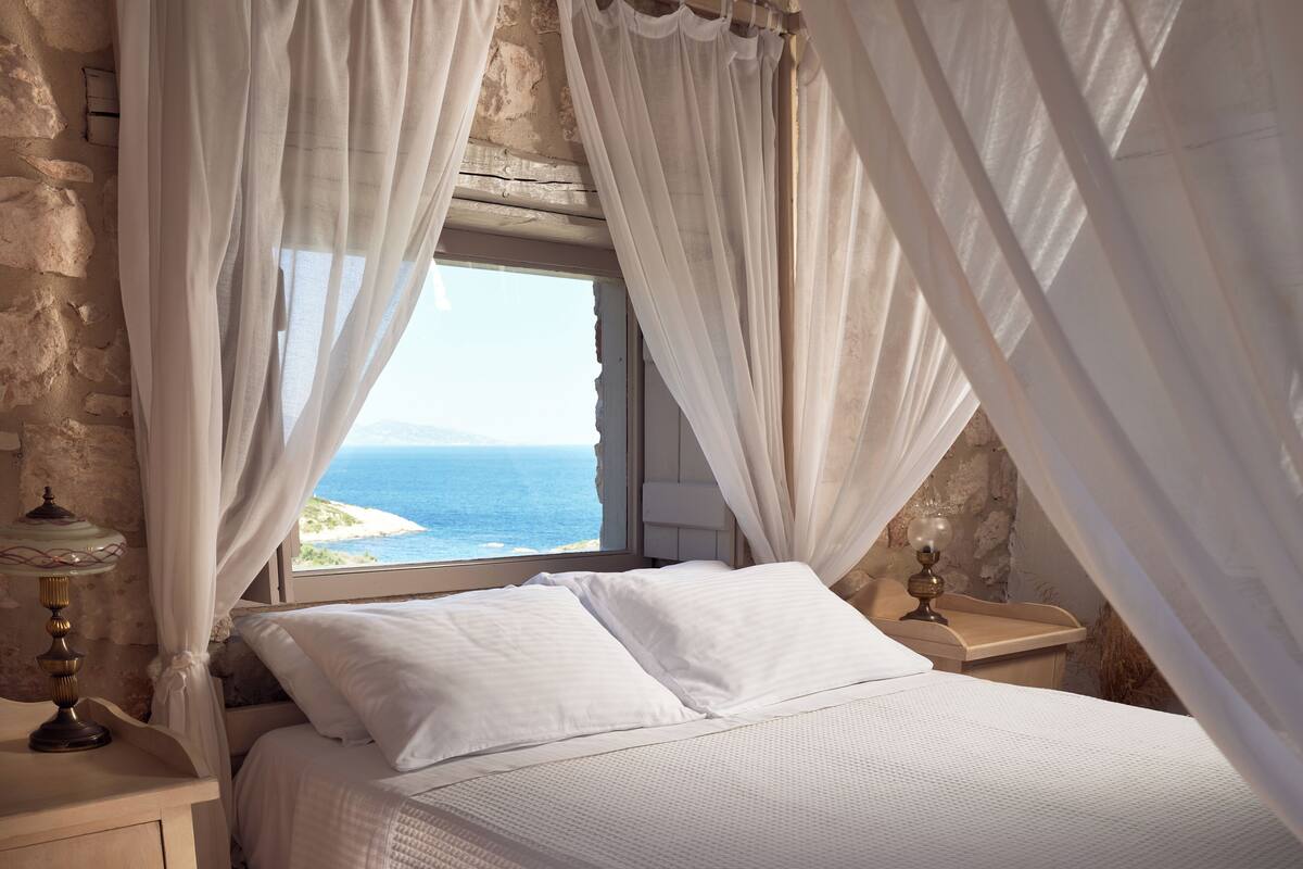 A bedroom with a bed, white curtains, a window with a view of the sea, and a lamp on a bedside table.