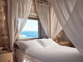 Un dormitorio con cama, cortinas blancas, una mesita de noche con lámpara y vista al mar a través de una ventana.