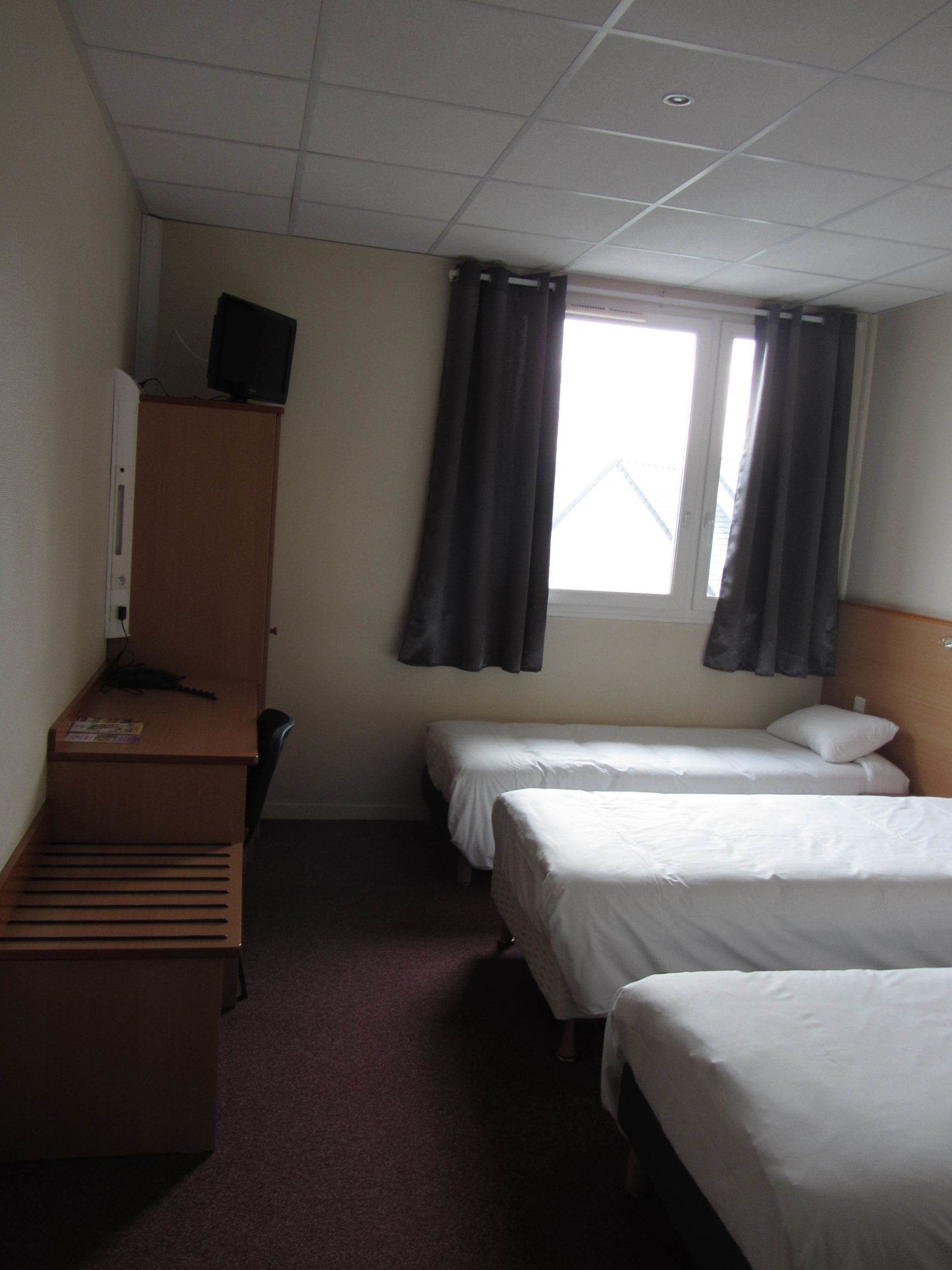 A hotel room with two single beds, a desk with a computer, and a window with curtains.