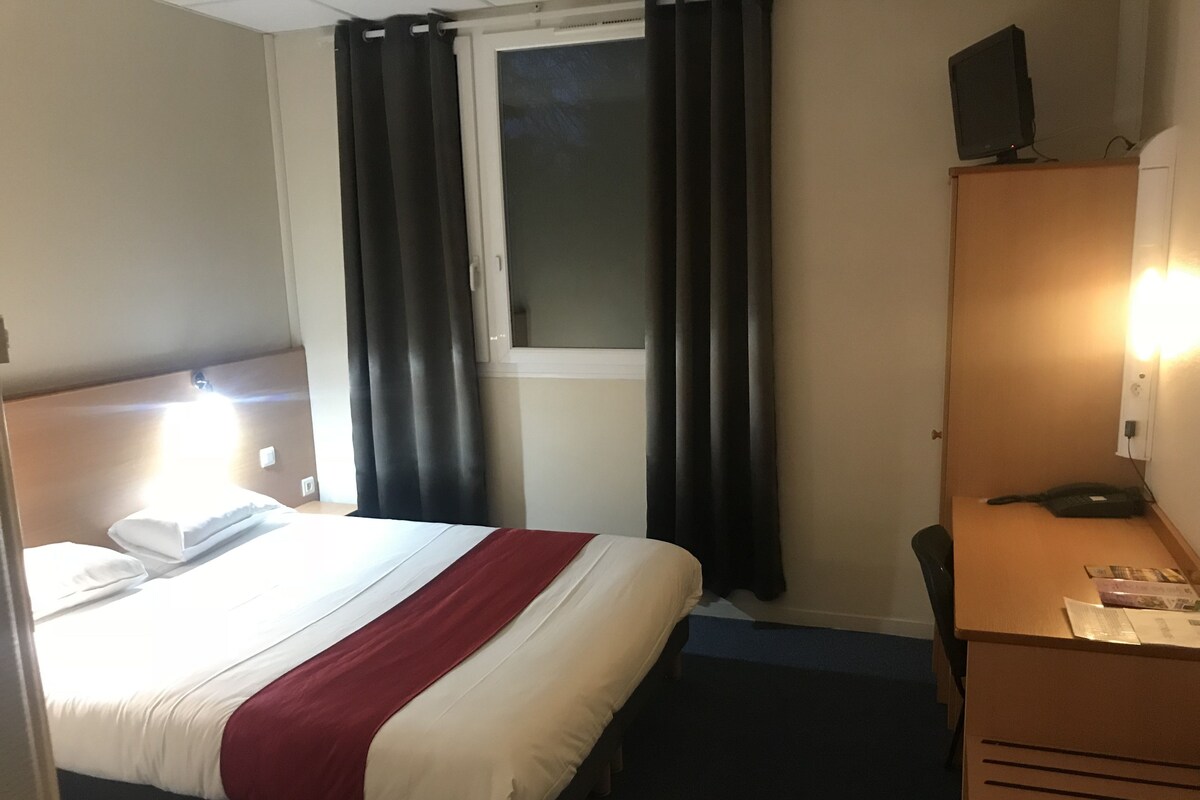 Une chambre d’hôtel comprenant un lit, un bureau, une chaise, une fenêtre avec des rideaux, un téléviseur posé sur un meuble et un lampadaire fixé au mur.