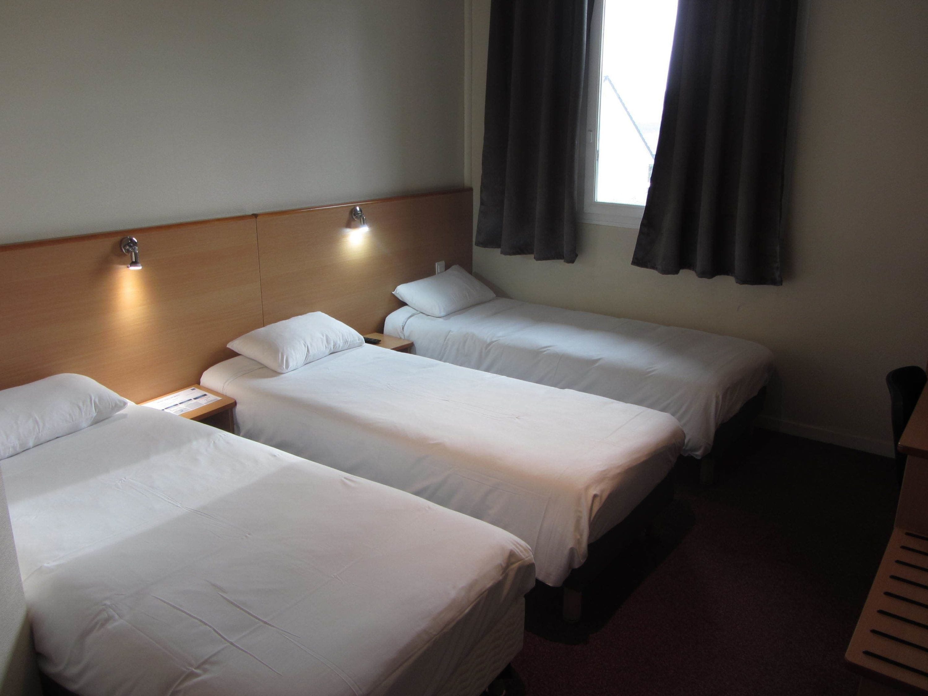 A hotel room with three single beds, a wooden headboard, a nightstand with a lamp, and a window with curtains.