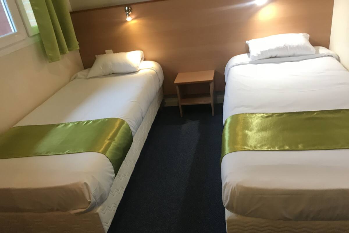 Two single beds with green bedspreads, a small wooden nightstand, and a window with green curtains.