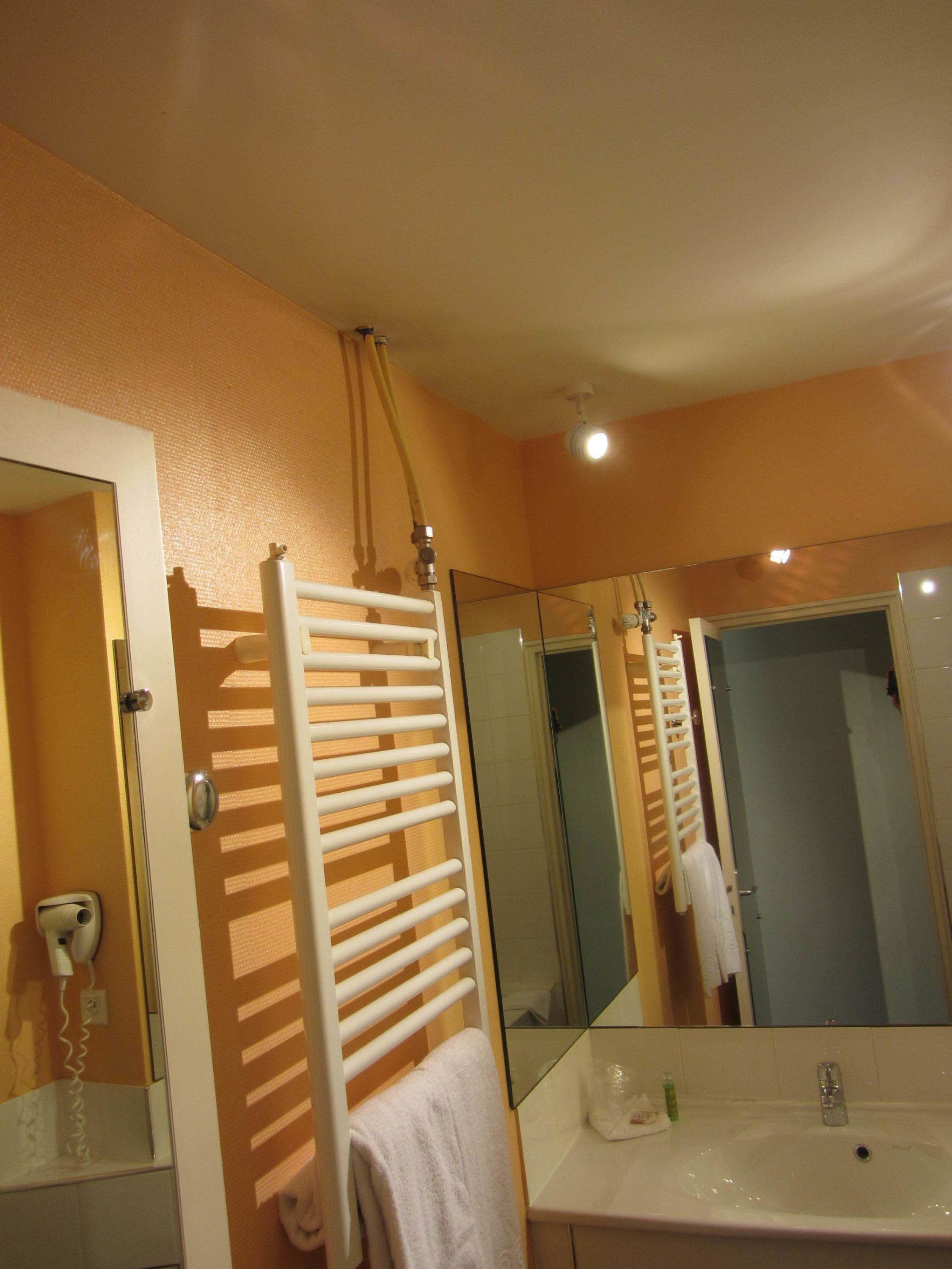 Une salle de bain avec un lavabo blanc, un miroir, un porte-serviettes et une douche visible dans le reflet.