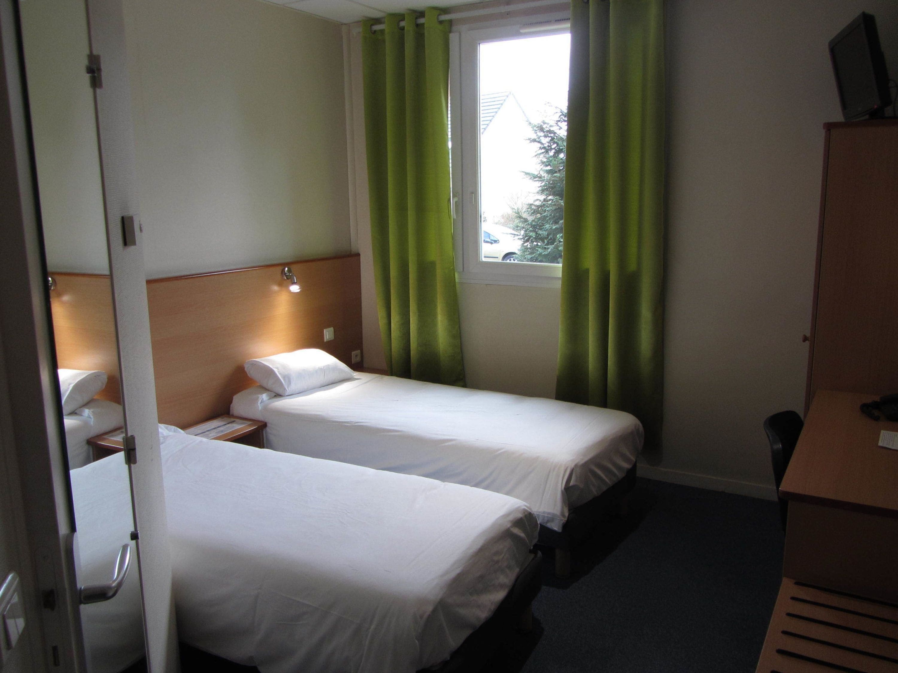 Ett hotellrum med två separata sängar, en sänggavel i trä, ett nattduksbord med en lampa och ett fönster med gröna gardiner.