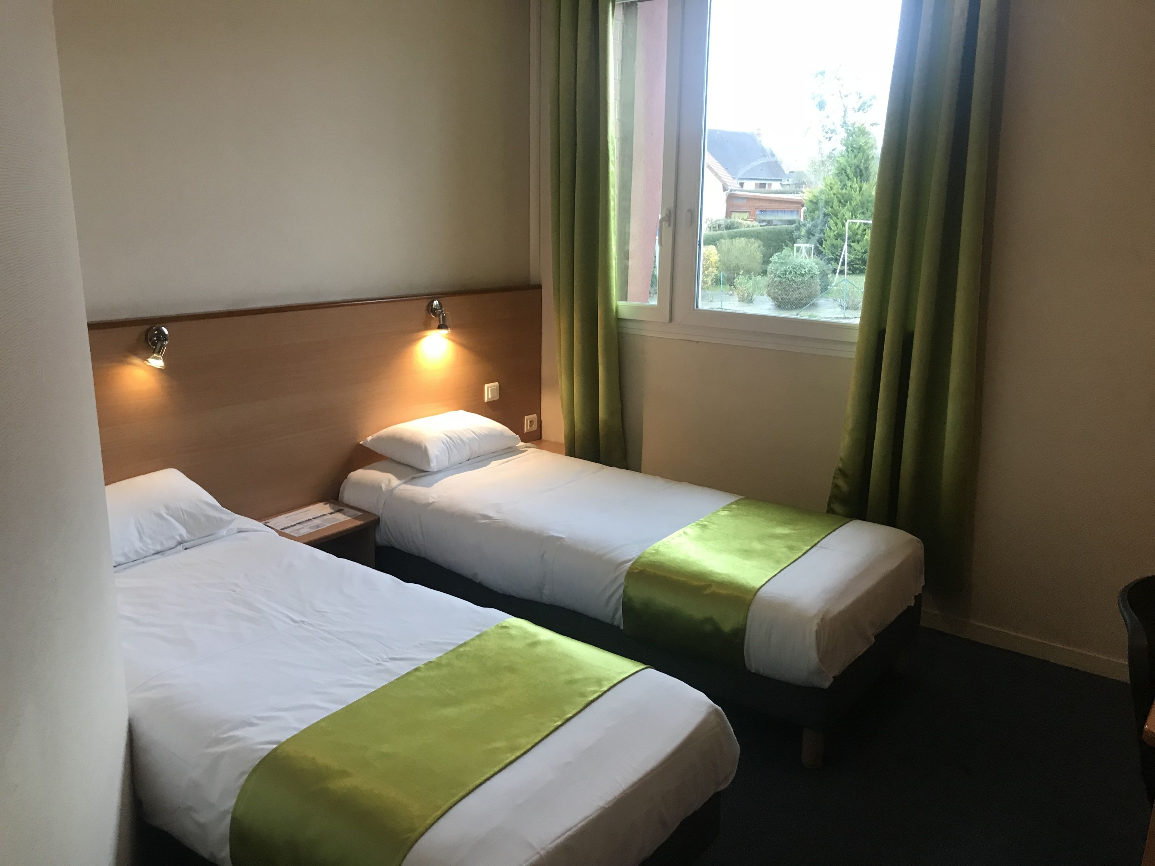A hotel room with two single beds, a nightstand, a window with curtains, and a view of the outside.