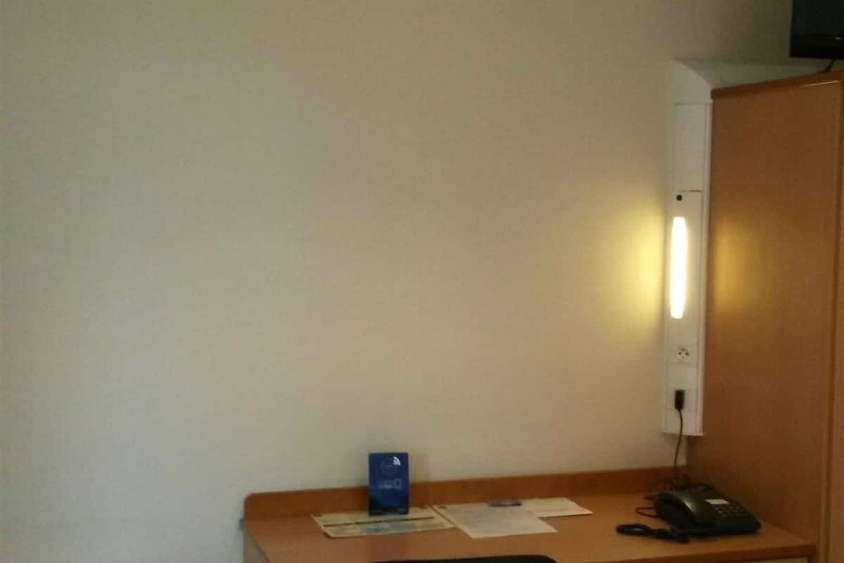 A hotel room with a wooden desk, a chair, a small trash bin, a bedside table, and a television on the wall.