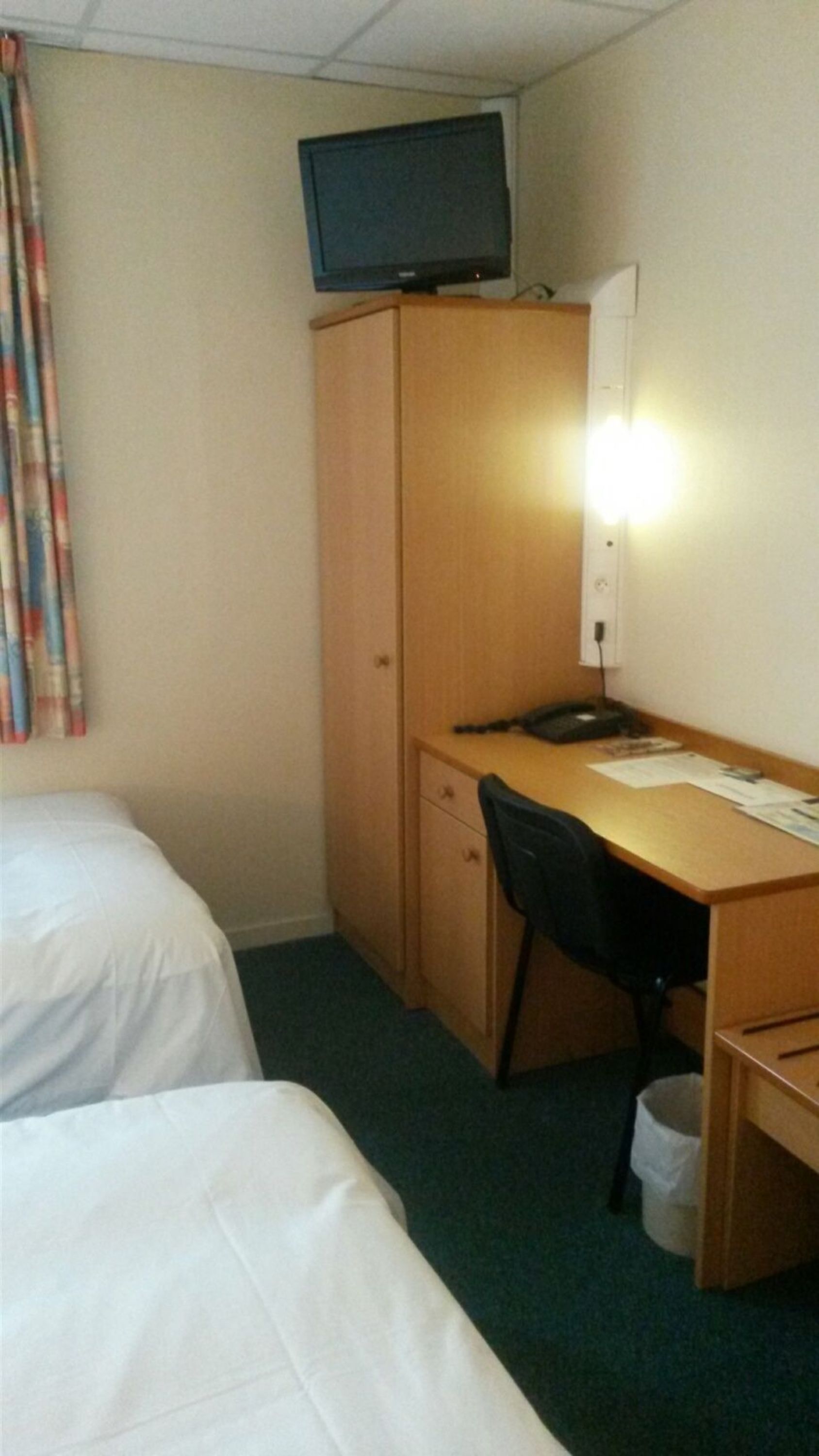 A hotel room with two single beds, a wooden desk, a chair, a television on a stand, and a desk lamp.