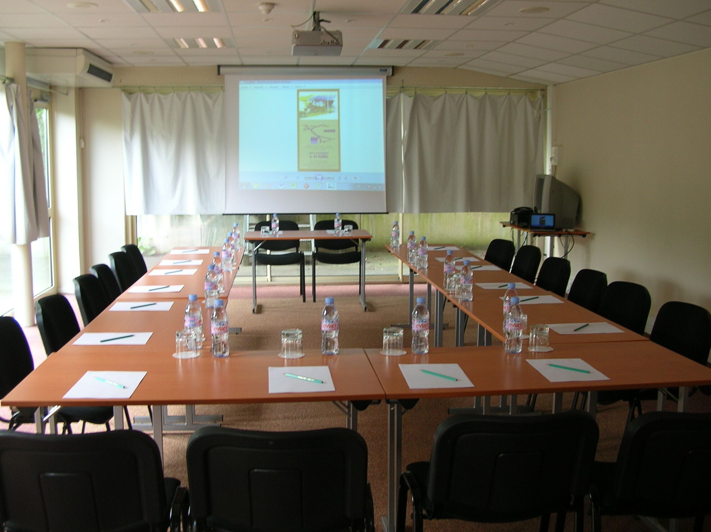 Ein Konferenzraum mit einem langen Tisch, Stühlen, Wasserflaschen und einer Projektionsleinwand, die eine Präsentation zeigt.