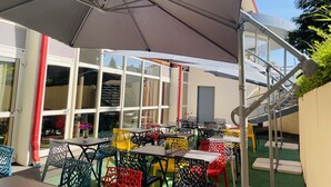 Un espace extérieur aménagé avec des tables et des chaises, un grand parasol et un escalier menant à un niveau supérieur.