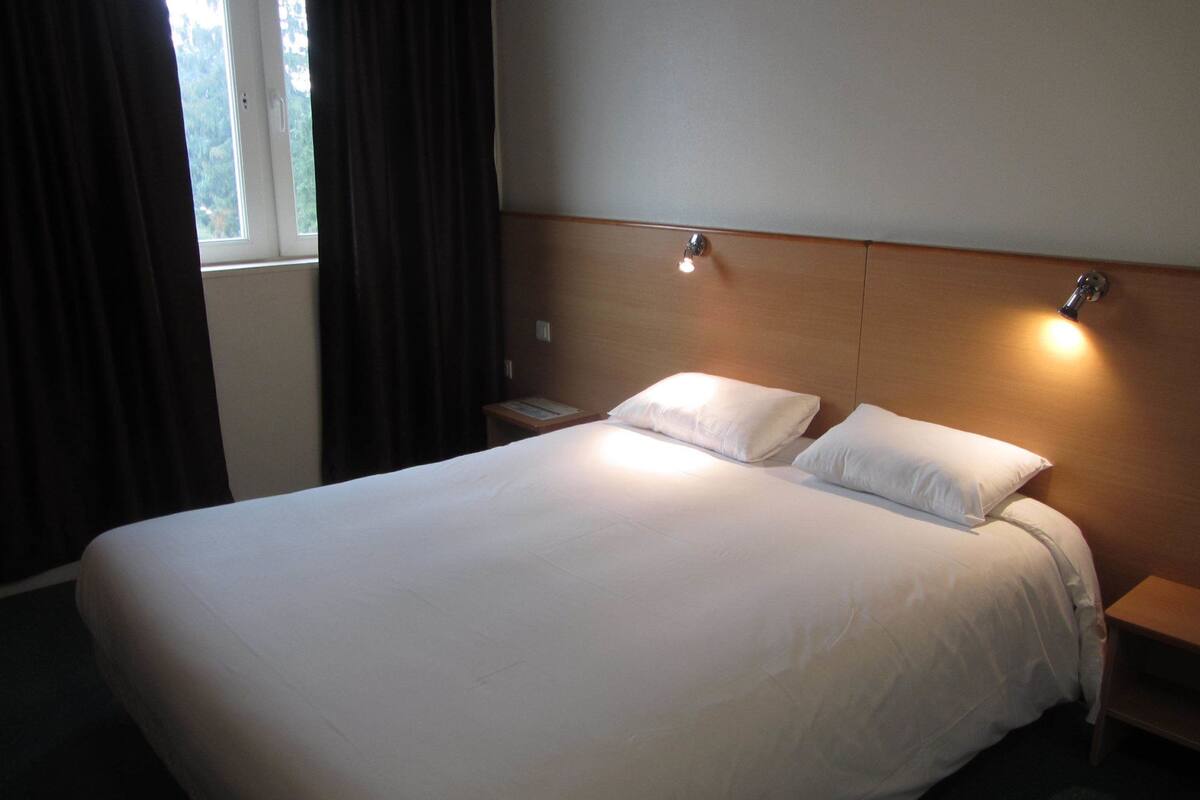 A hotel room with a large bed, two bedside tables, a nightstand, and a window with curtains.
