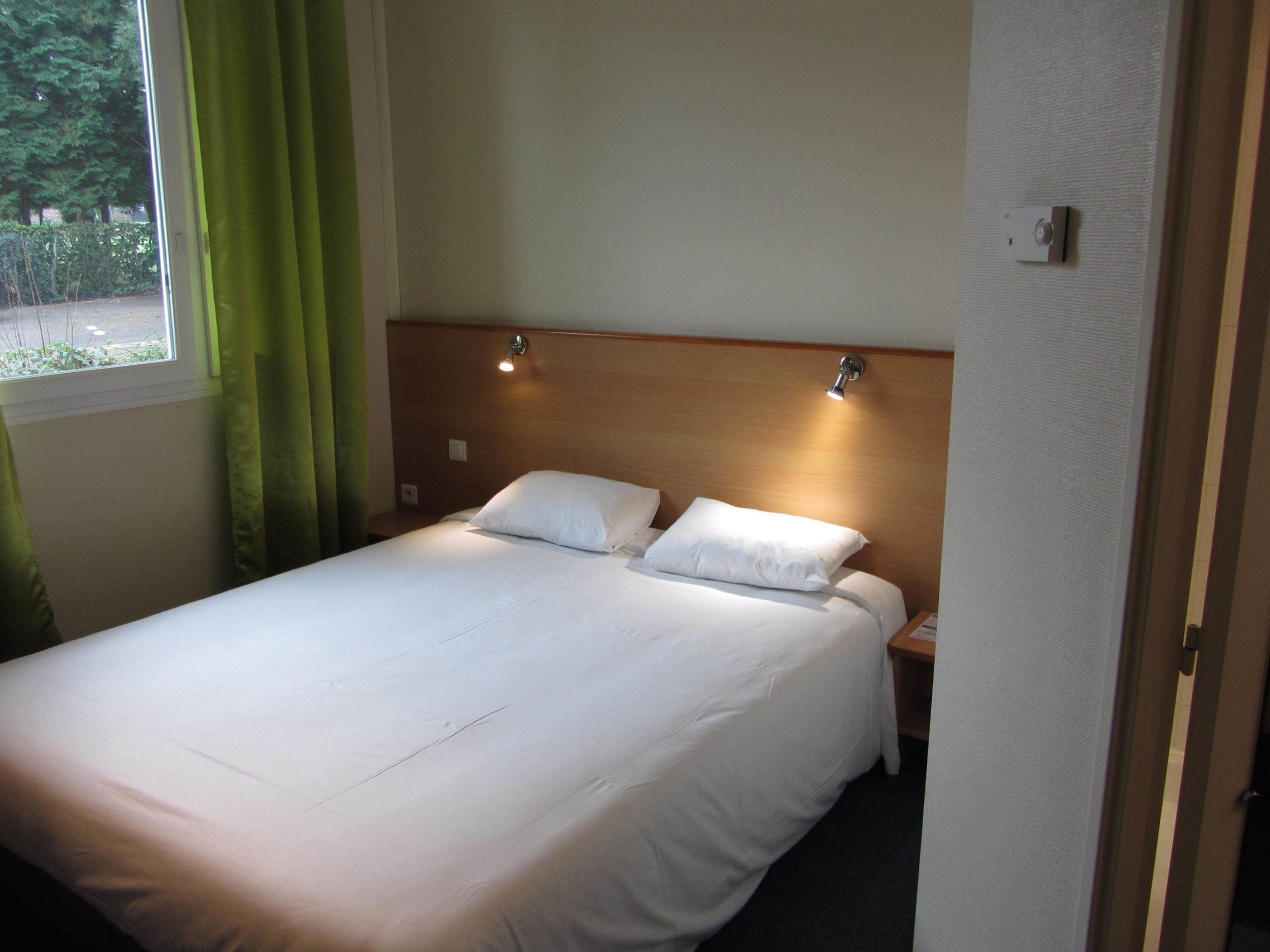 Una cama bien hecha con dos almohadas en una habitación de hotel con cabecera de madera, una mesita de noche y una ventana con cortinas.