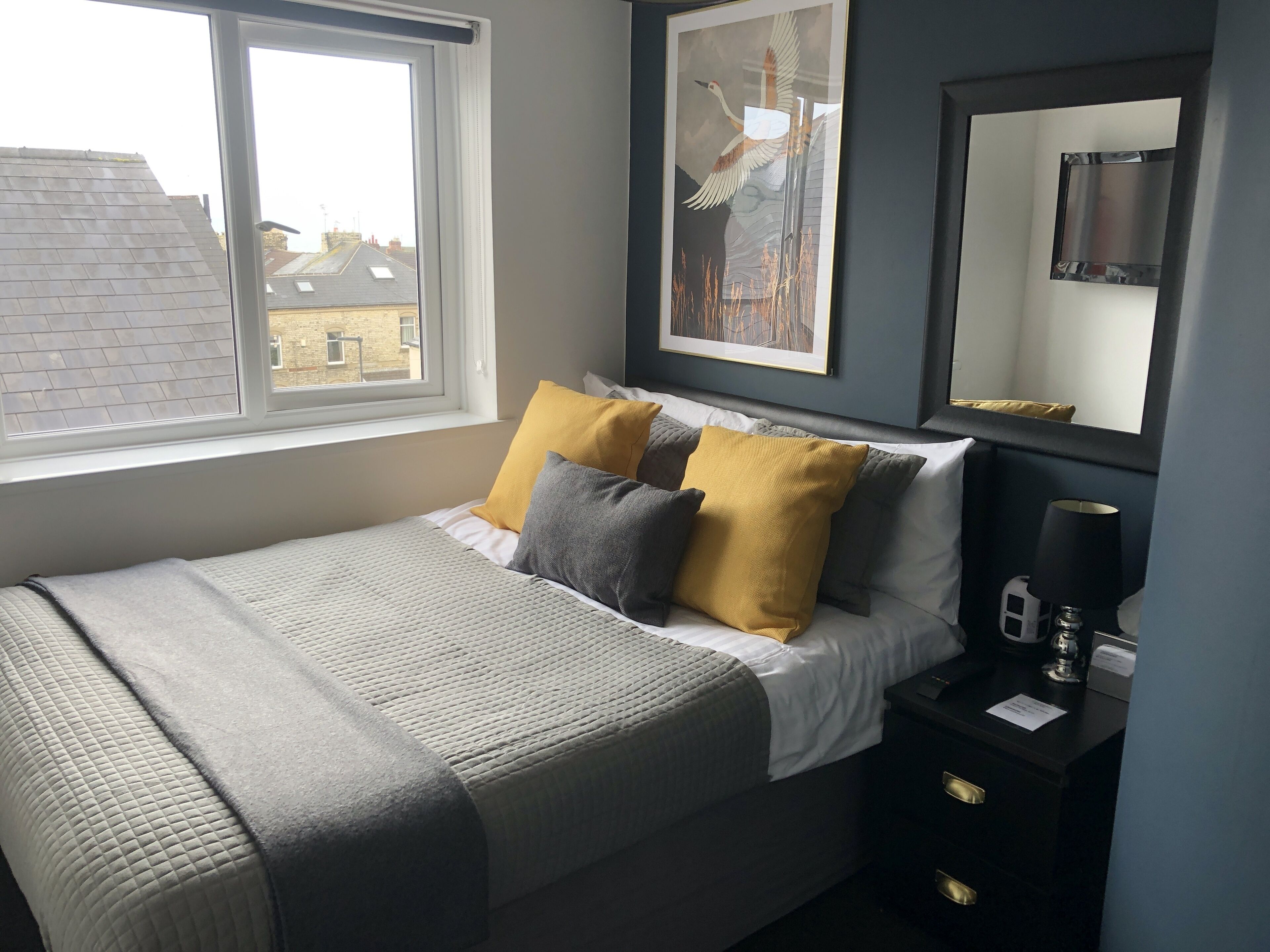 A neatly made bed with a grey and white duvet, two pillows in yellow and grey, a bedside table with a lamp, and a framed artwork above the bed.