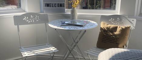 A room with a white table, two white chairs, a 'WELCOME' sign, a vase with yellow flowers, and a view of a building with arches.