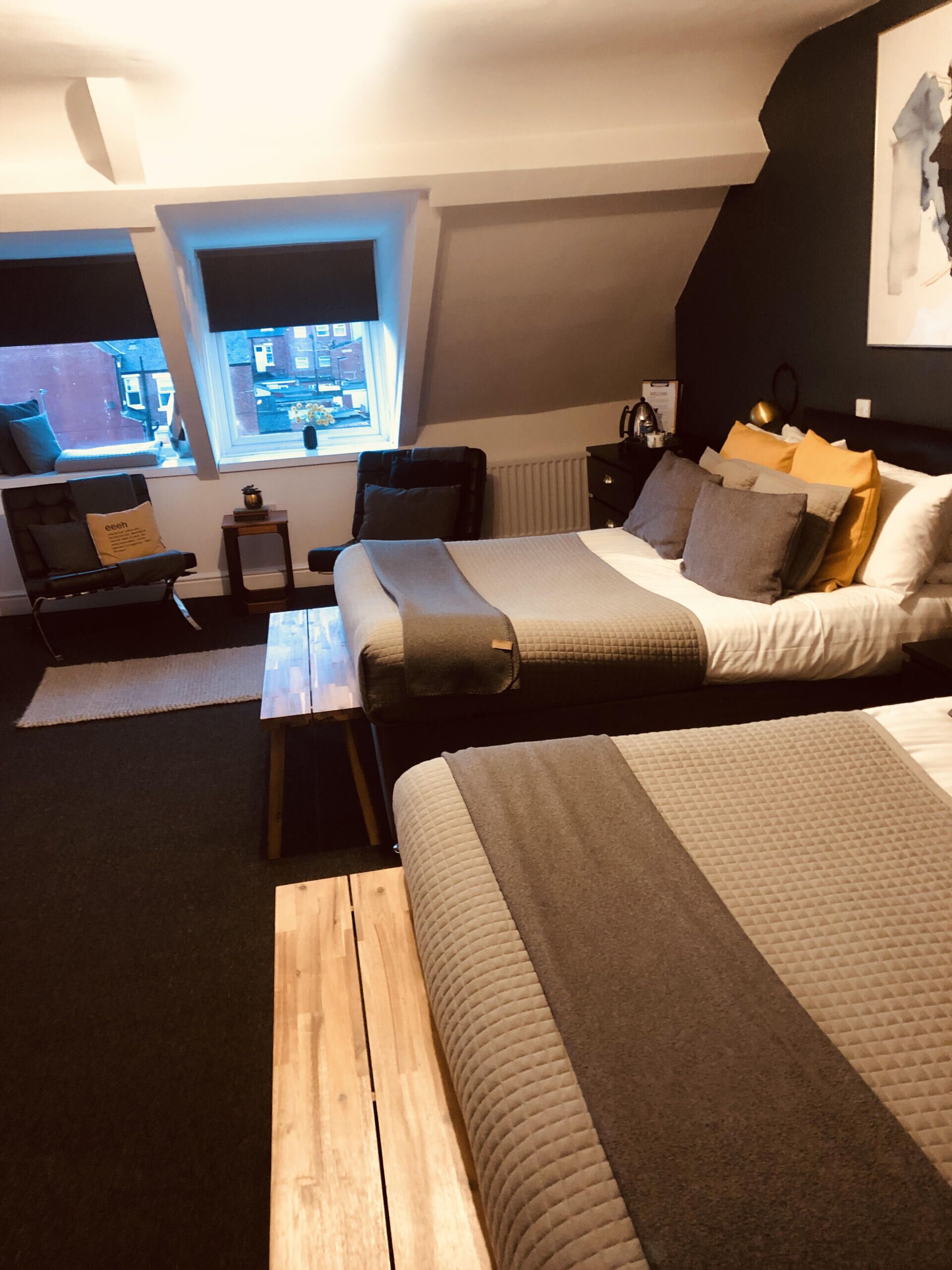 A modern bedroom with a large bed, a small sofa, a wooden table, and a view of the city through a window.