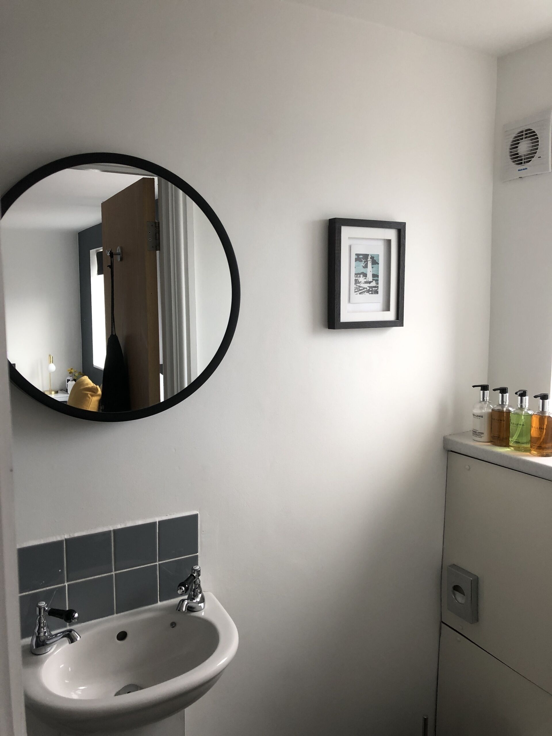 A bathroom with a round mirror, a framed picture, a sink, and toiletries on a cabinet.