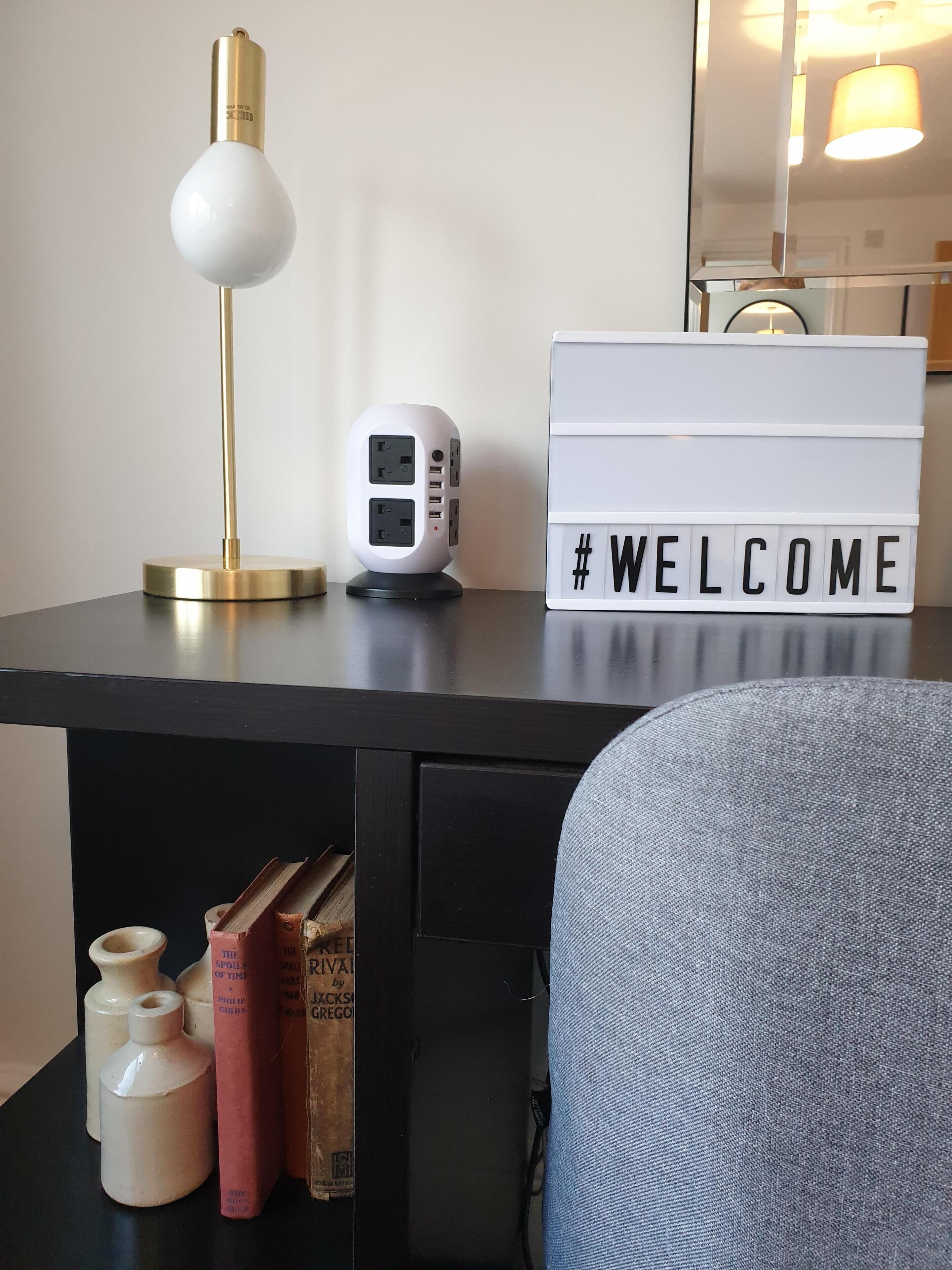 Un bureau avec une lampe, une multiprise, un panneau d’accueil et des livres.