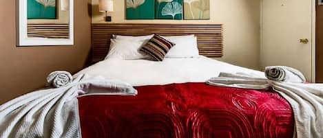 A hotel room with a bed, a red blanket, two pillows, a headboard, and artwork on the wall.