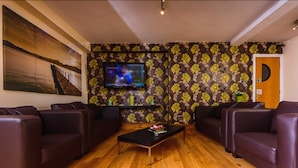 Un salón con suelo de madera, sofás de cuero oscuro, una mesa de centro negra, un televisor montado en la pared y un estampado de flores en el papel tapiz.