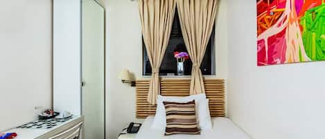 Une chambre d’hôtel avec un lit, un bureau, une chaise, une fenêtre avec des rideaux et un tableau coloré au mur.