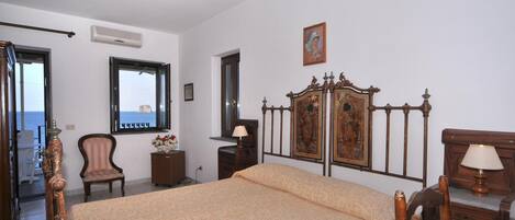 A bedroom with a large bed, a wooden headboard, a ceiling fan, a window with a view, and a small table with a lamp.