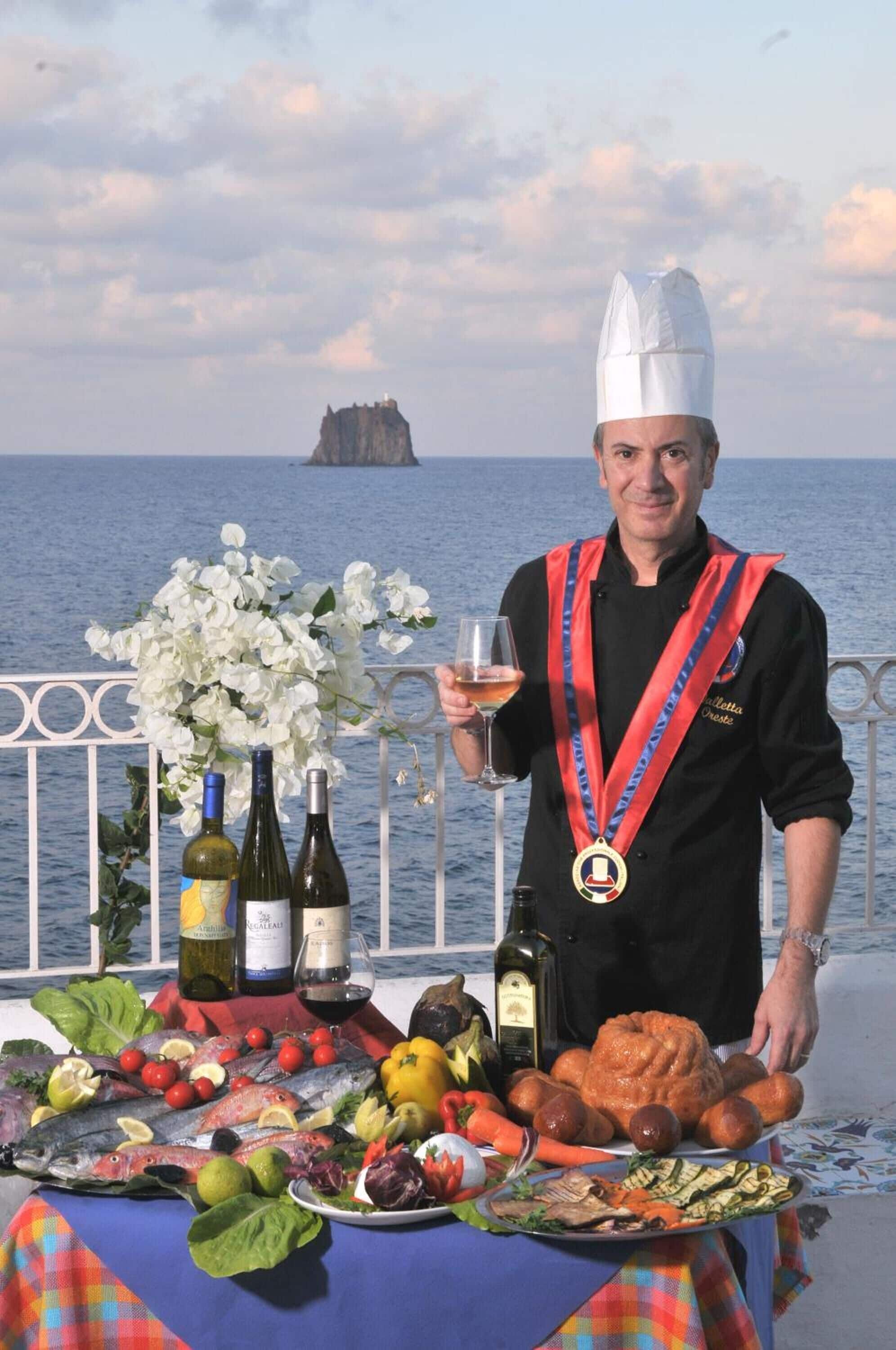A chef holding a glass of wine with a medal, standing by a table with seafood, vegetables, and wine bottles.