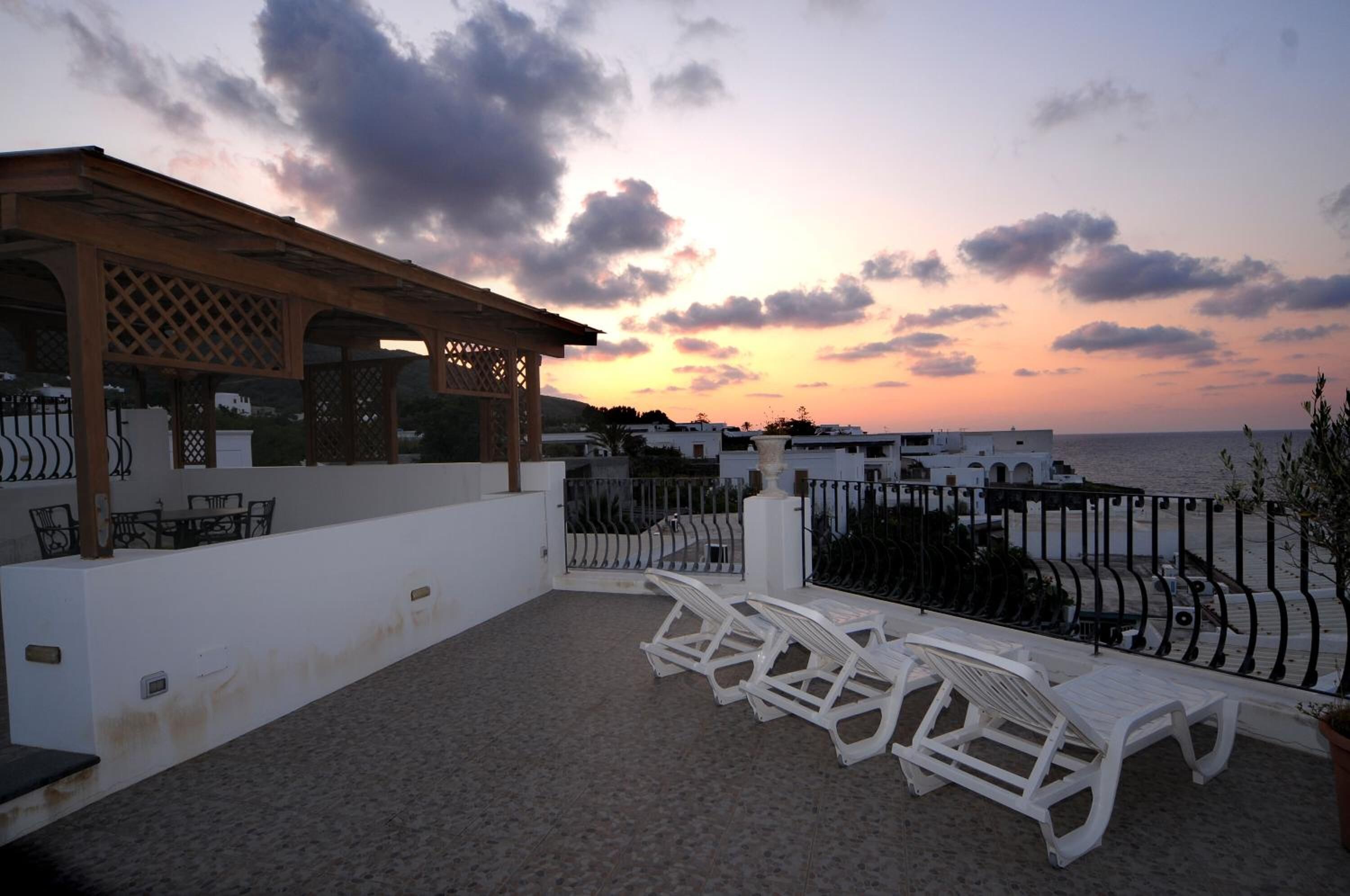 A terrace with white lounge chairs, a pergola, and a view of the sea at sunset.