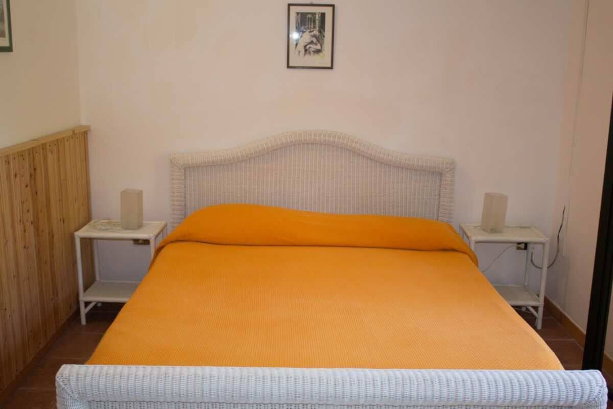 Un letto singolo con copriletto arancione, due comodini con lampade, un pannello in legno a sinistra e un quadro appeso al muro.