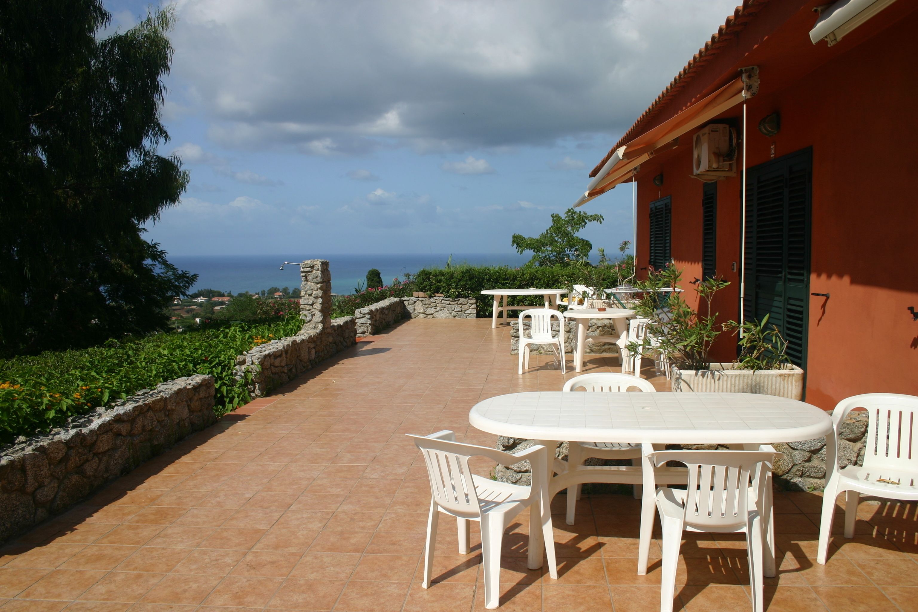 An outdoor patio with white plastic furniture, a stone wall, and a view of the sea.