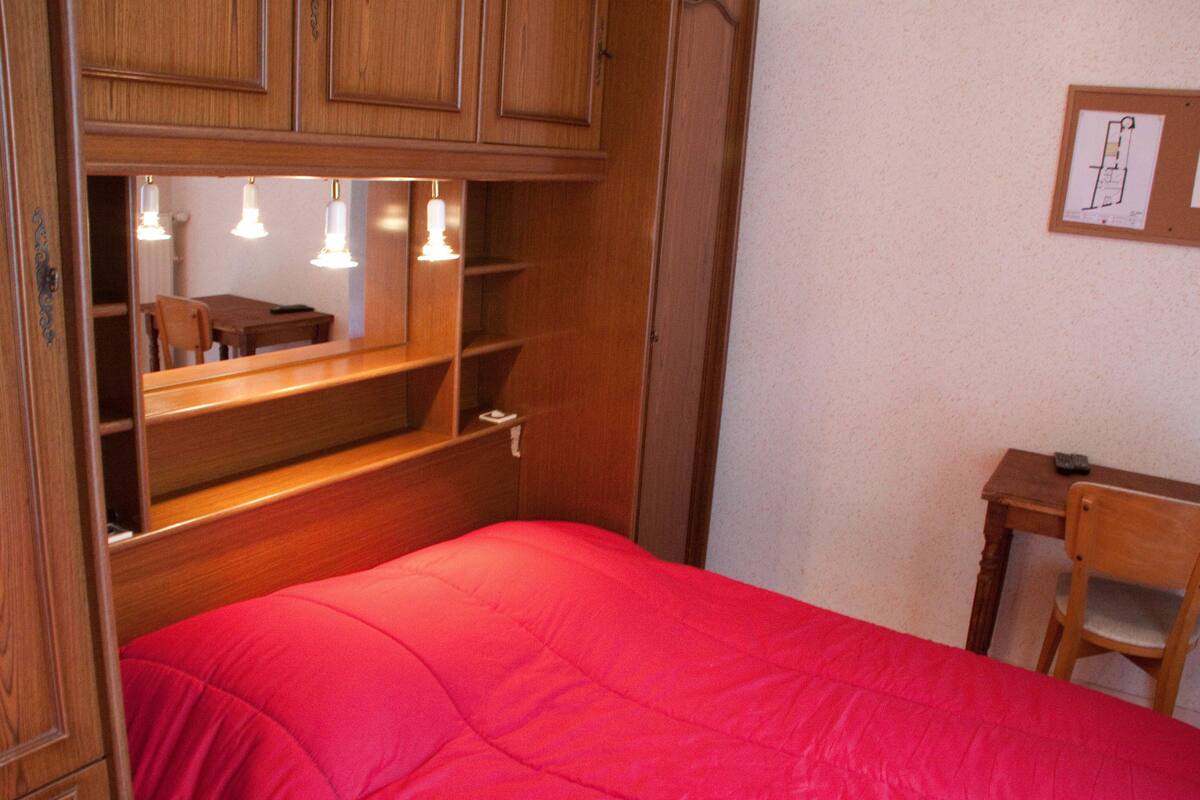 A bedroom with a red bedspread, wooden wardrobe, and a desk with a chair.