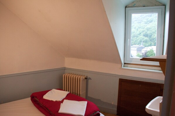 A small, narrow room with a single bed, a radiator, a window with a view of buildings, and a wooden cabinet.