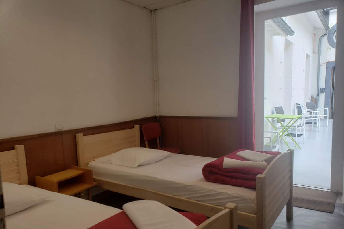 A hotel room with two single beds, a wooden nightstand, a red chair, and a view of the outdoor seating area through a glass door.
