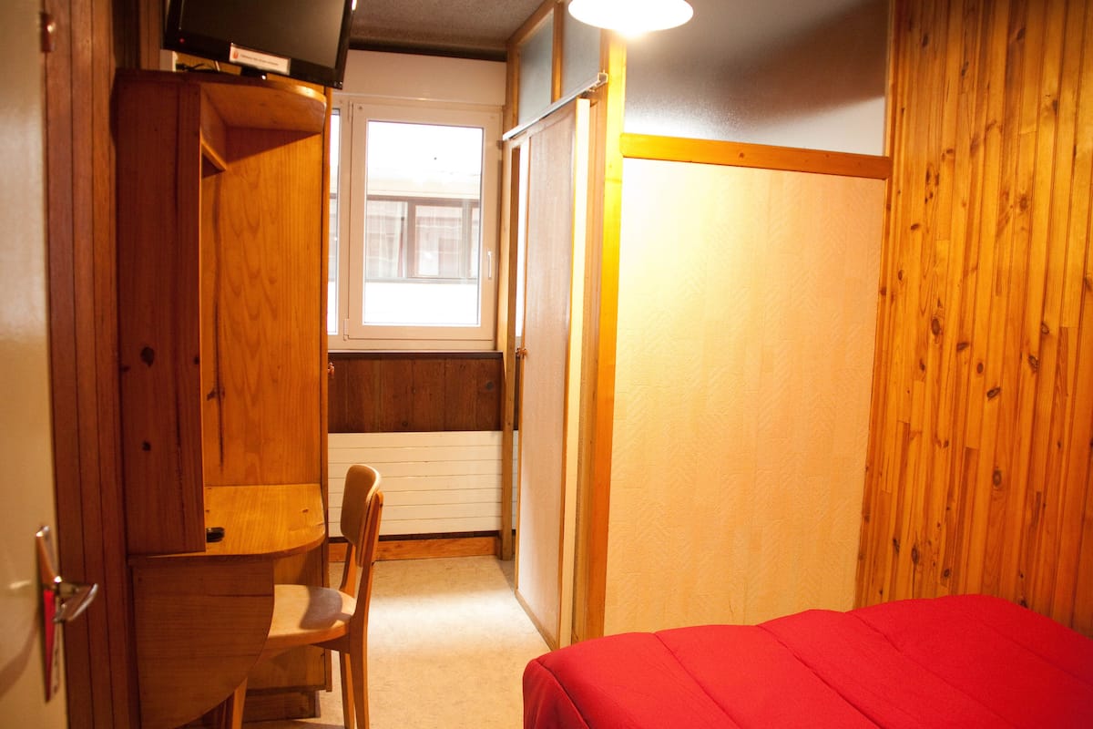 A bedroom with a wooden wardrobe, a chair, a bed with a red cover, and a window with blinds.