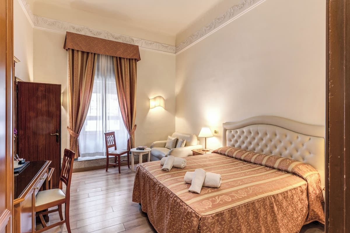 A hotel room with a bed, a desk, a chair, a lamp, and a window with curtains.