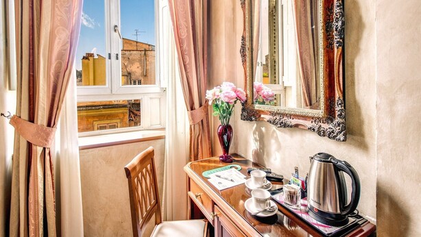 A hotel room with a desk, a chair, a mirror, a vase with flowers, a kettle, and a window with curtains. - Hotel Labelle  (Rome)