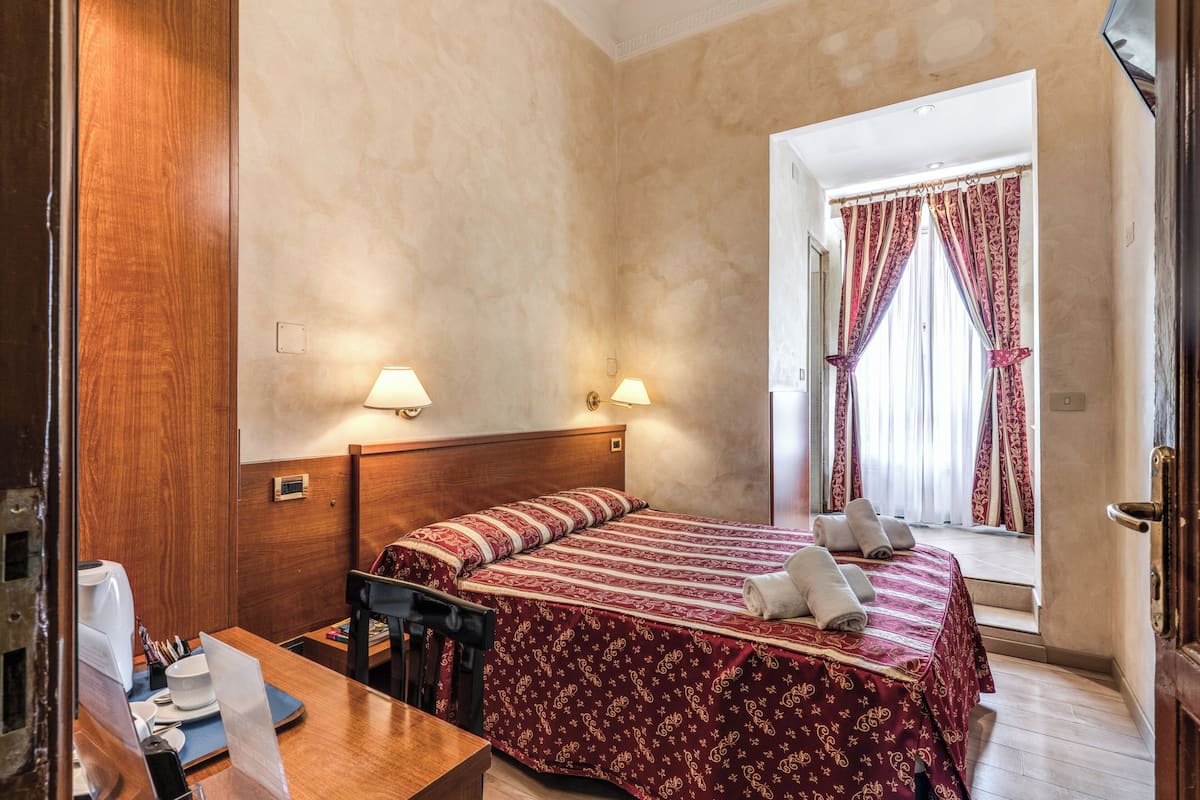 A hotel room with a wooden headboard, two bedside lamps, a bed with a patterned bedspread, a desk with a chair, and a window with curtains.