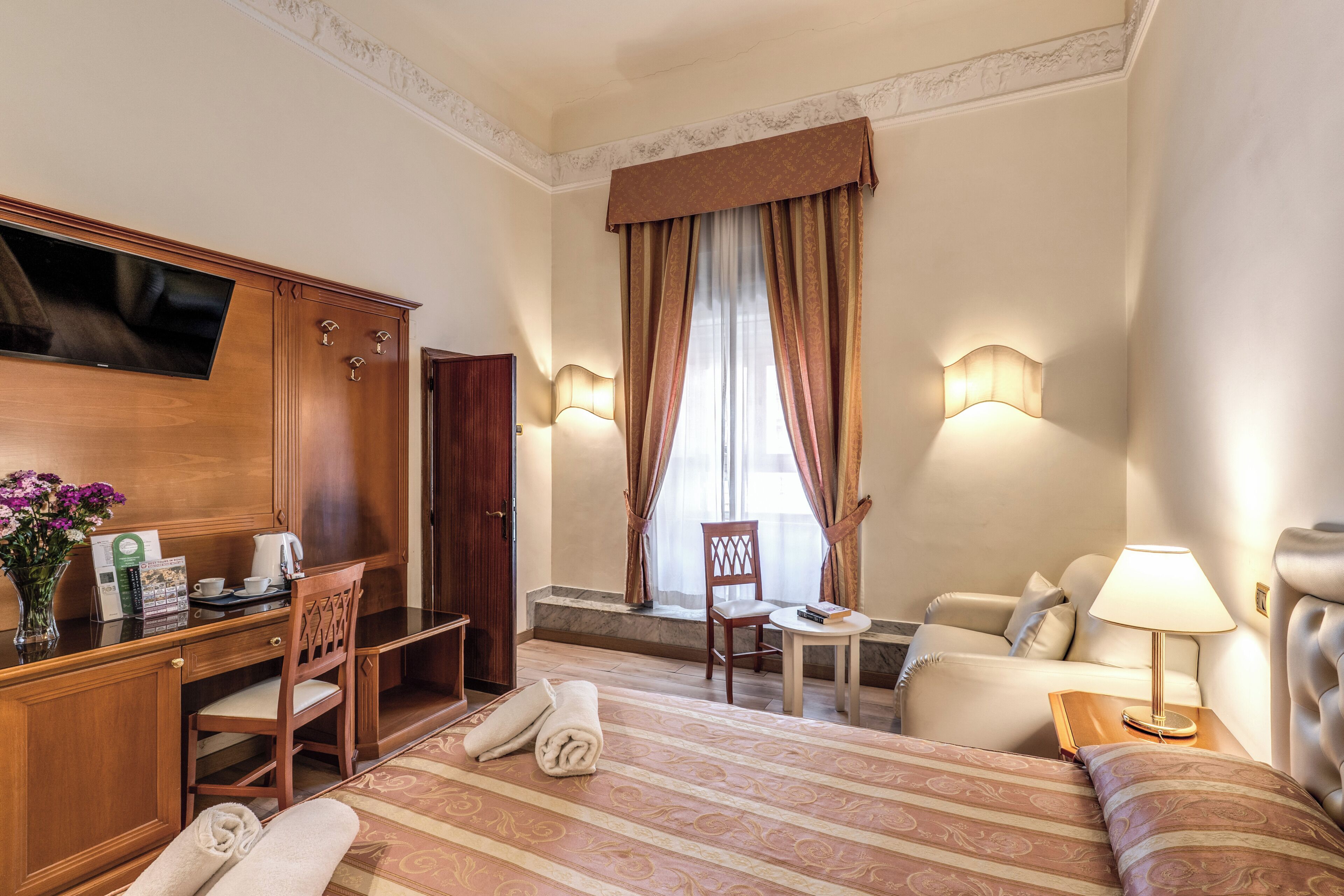 Ein Hotelzimmer mit einem Bett, einem Schreibtisch mit Fernseher, einem Sessel, einem kleinen Tisch, einer Lampe und einem Fenster mit Vorhängen.