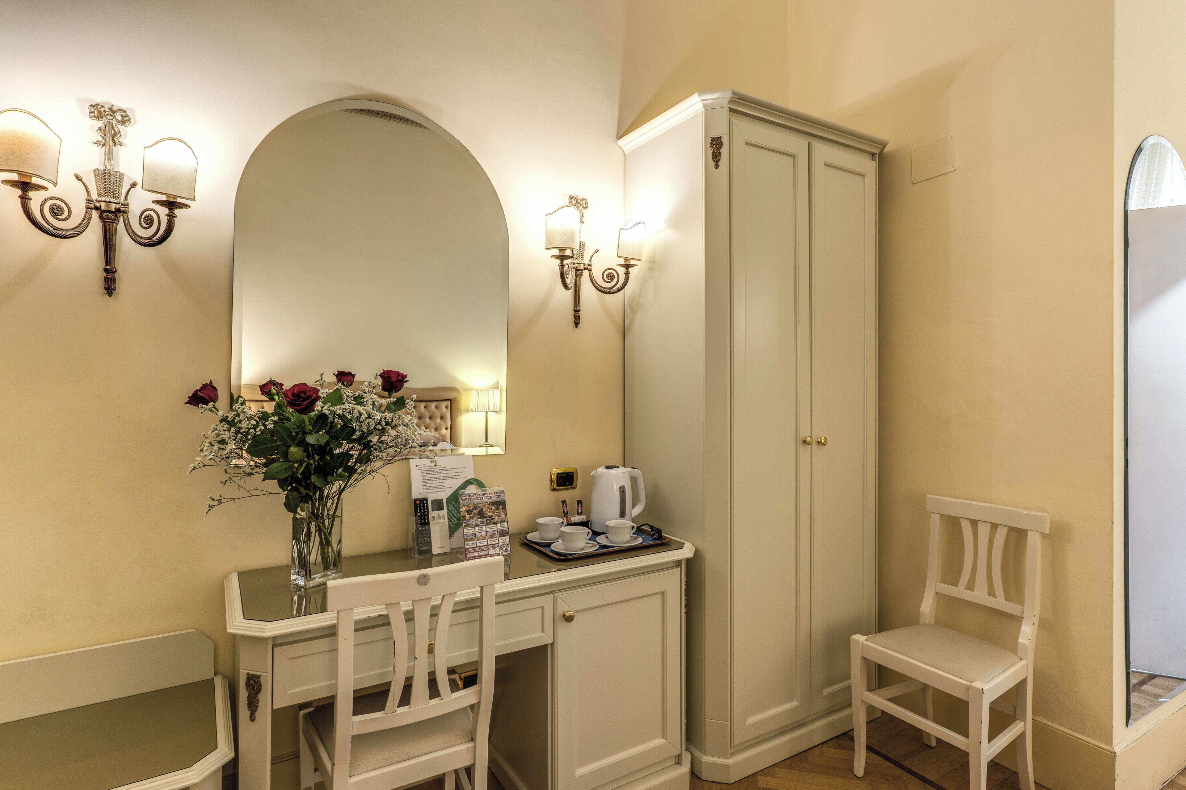 A room with a vanity, a mirror, a cabinet, a chair, and a vase with flowers.