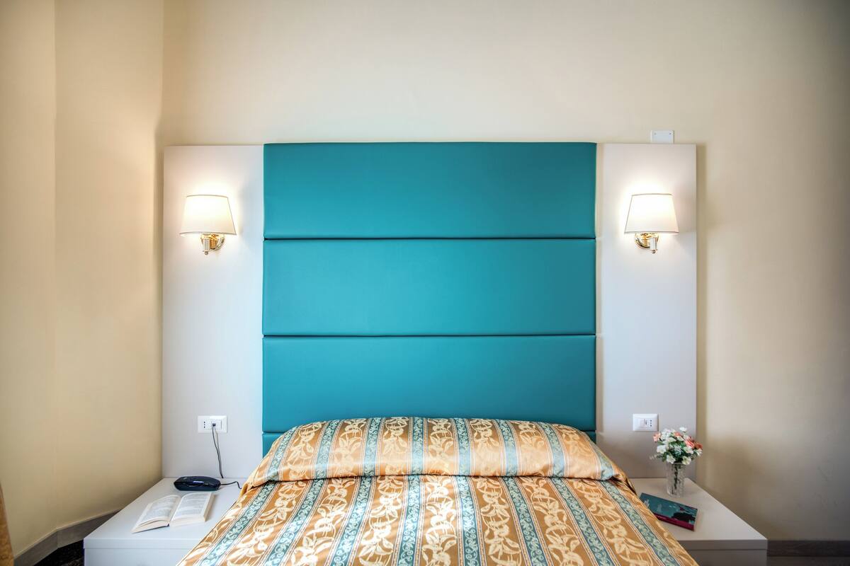 A hotel room with a teal headboard, two bedside lamps, a nightstand with a book, and a vase with flowers.