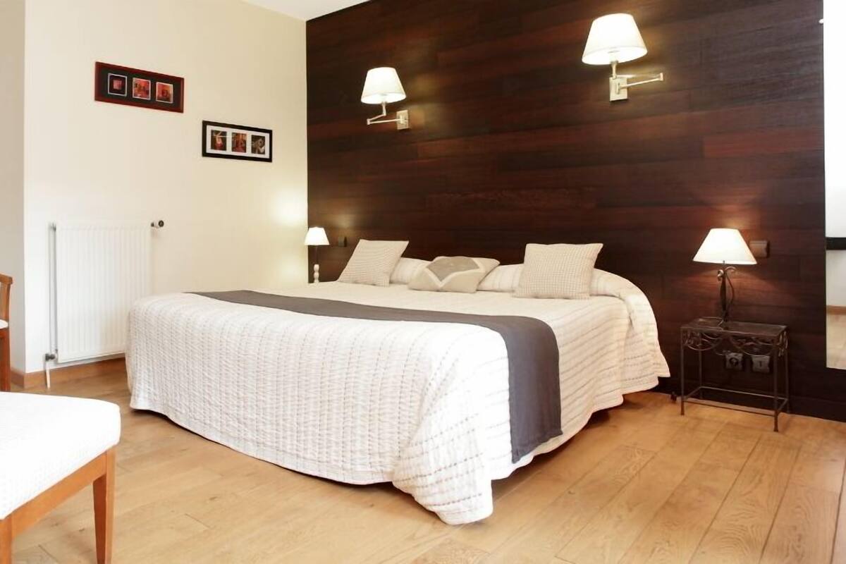A bedroom with a bed, bedside lamps, a wooden headboard, a desk with a chair, and framed pictures on the wall.
