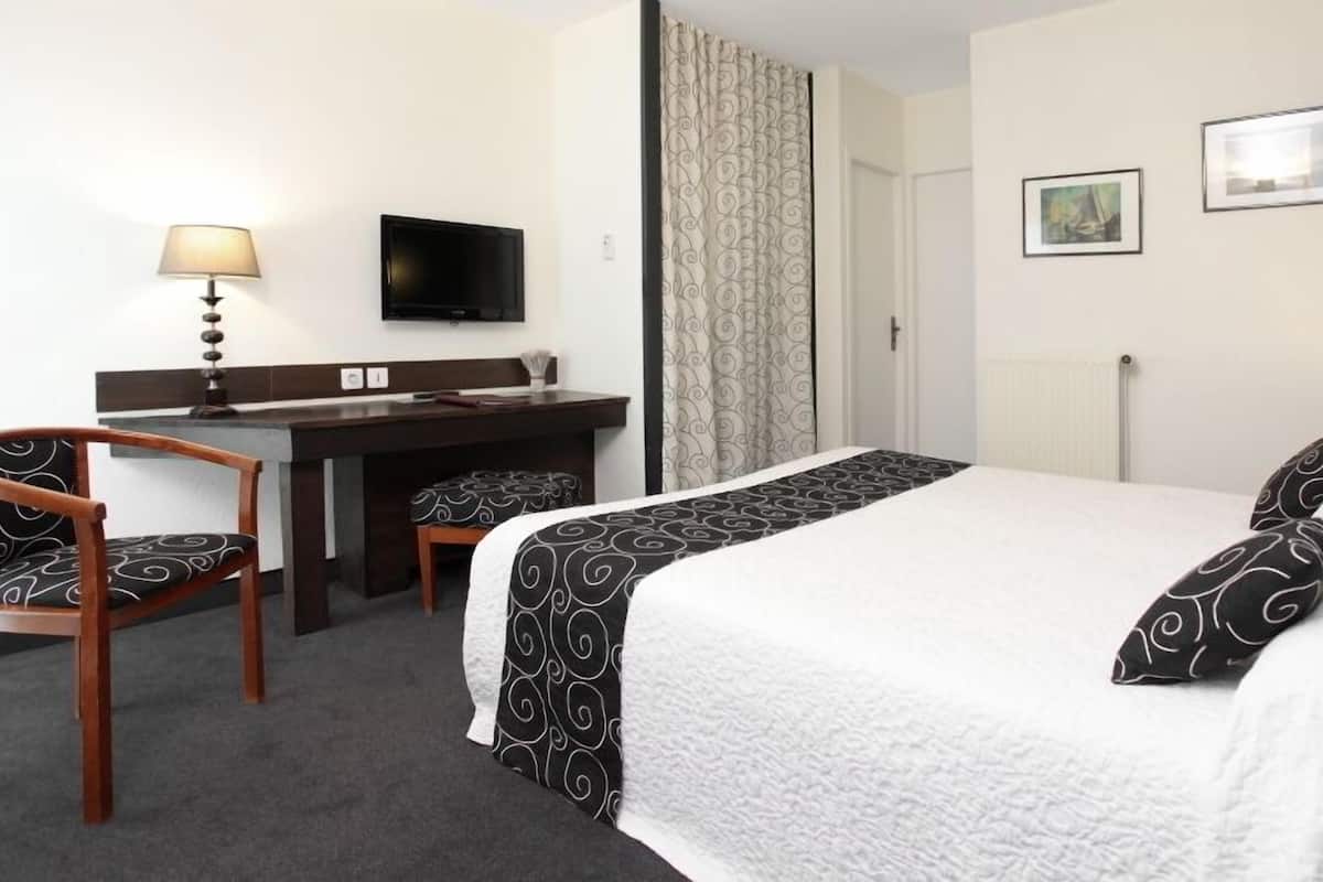 A hotel room with a bed, a desk, a chair, a TV, and two framed pictures on the wall.