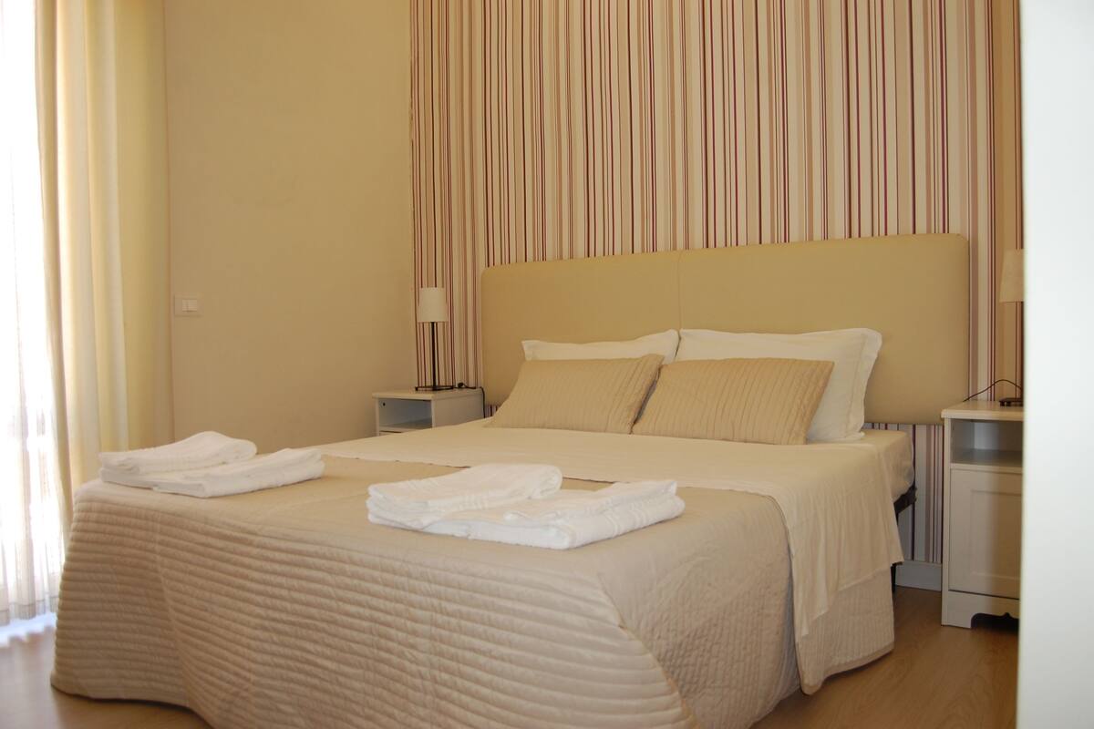 Una cama bien hecha con toallas dobladas, una lámpara de noche y una cortina a rayas en una habitación de hotel.
