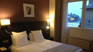 A hotel room with a large bed, two bedside lamps, a nightstand with a phone, and a window with a view of buildings. - Hotel de la Plage (Dinard)