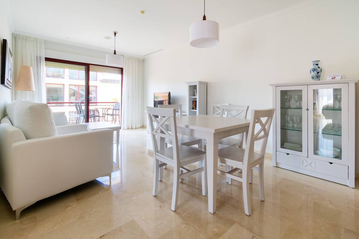 A modern living room with a white sofa, dining table, and chairs, a cabinet with glass doors, and a large window with curtains.