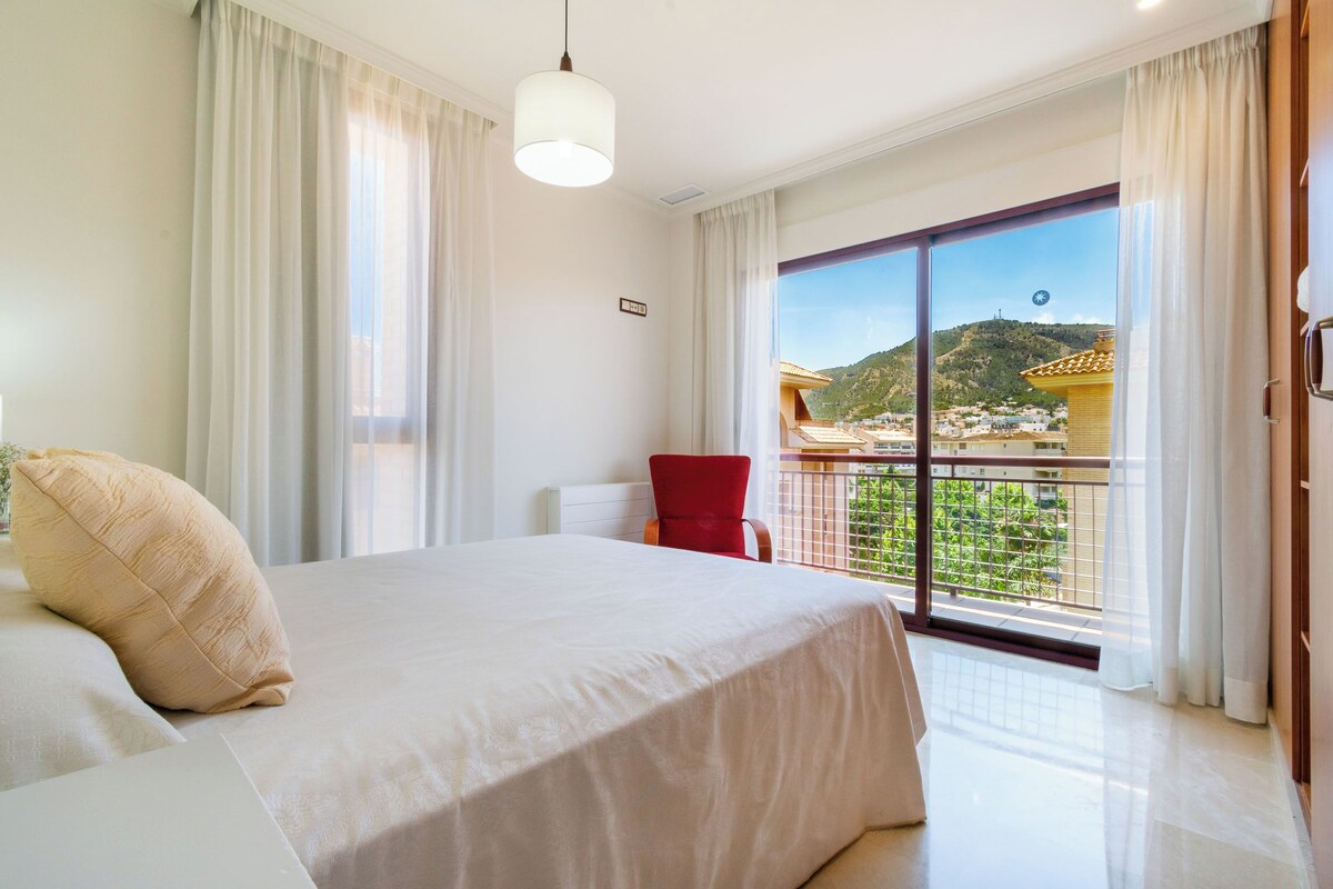 Habitación de hotel con una cama grande, una silla roja y un balcón que ofrece vistas a una colina y edificios.